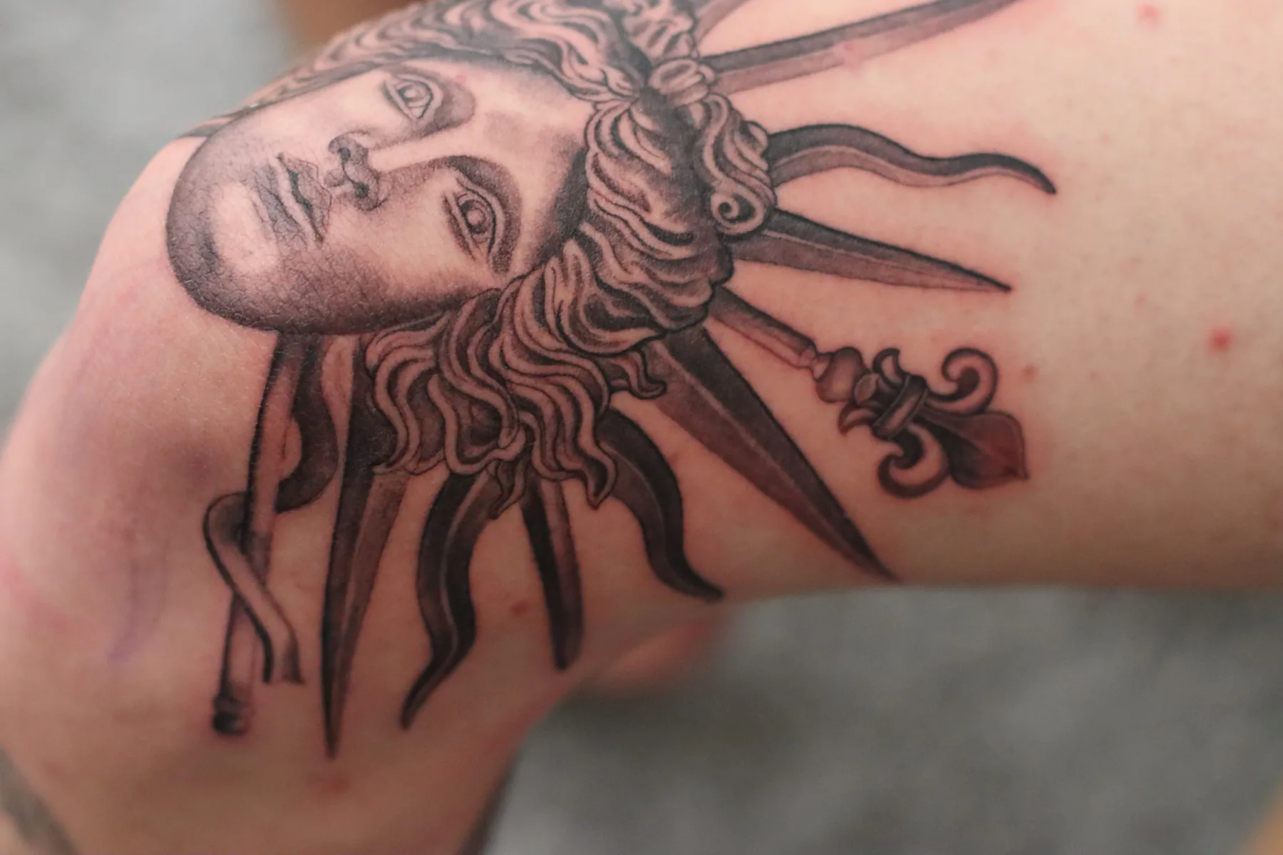 Black & Grey Tattoo eines antiken Gesichts mit lockigem Haar, Strahlenkranz und Lilienspitzen am Arm nahe dem Ellbogen, Wien