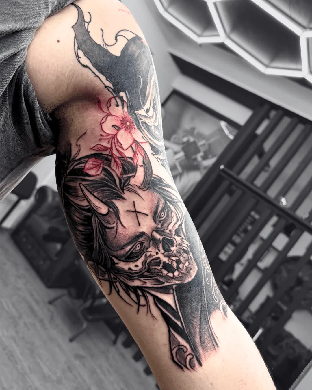Black & Grey Tattoo am Arm: Dämonin mit Horn, halbes Gesicht als Totenkopf, Kreuz auf der Stirn und rote Blüte - Tattoo Wien