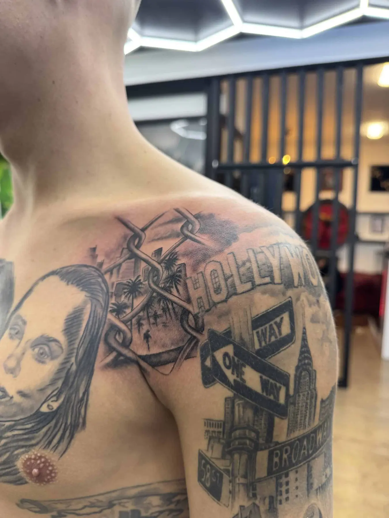Black & Grey Collage-Tattoo an der Schulter: Zaun mit Palmen, Hollywood-Schriftzug, Straßenschilder und Skyline, Wien