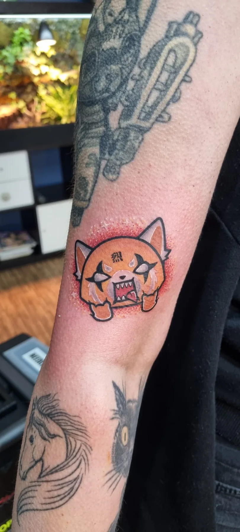Farbiges Cartoon Tattoo des wütenden roten Pandas Aggretsuko am Unterarm, zwischen älteren Tattoos, Tattoo Studio Wien