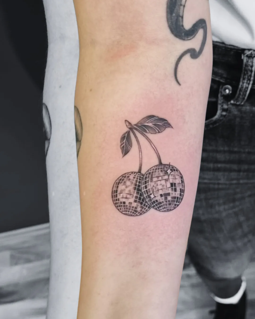 Fineline Tattoo zweier Kirschen als glitzernde Discokugeln mit Blättern am Unterarm, Black & Grey, Tattoo Studio Wien