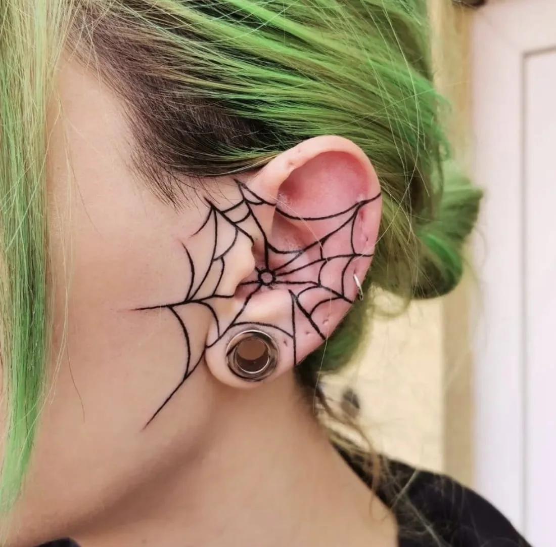 Fineline Spinnennetz-Tattoo über Ohrmuschel und Wange in Schwarz, neben einem Ohr-Tunnel, Tattoo Studio Wien