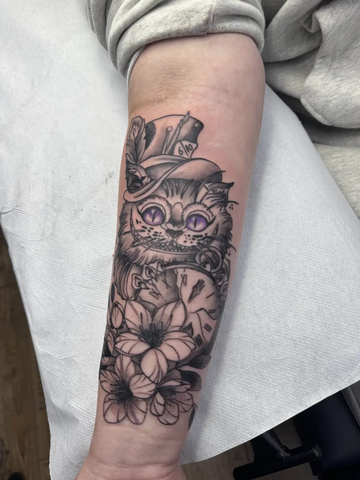 Tattoo der Grinsekatze aus Alice im Wunderland mit Zylinder, violetten Augen, Taschenuhr und Lilien am Unterarm, Wien