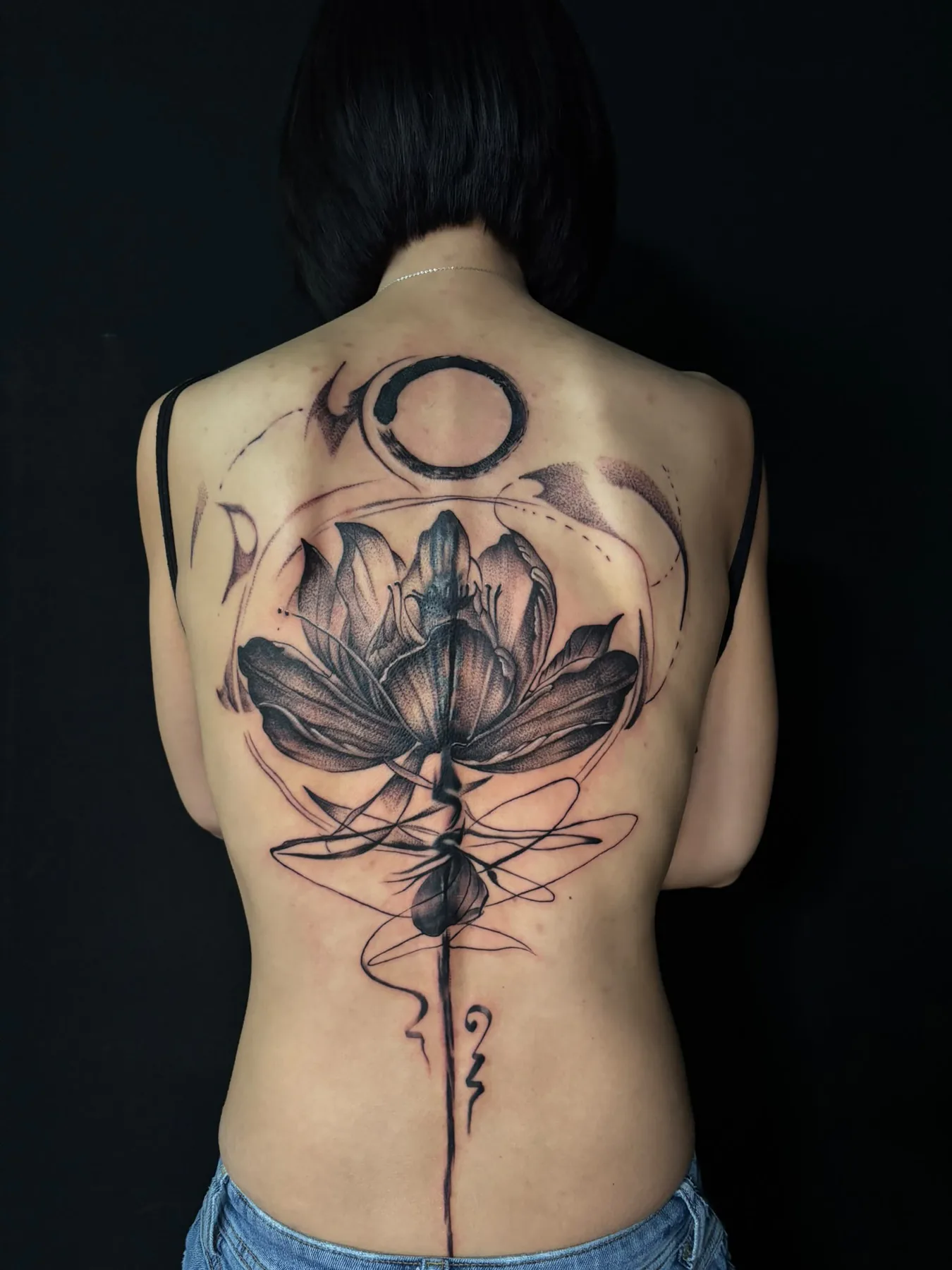 Großes Rückentattoo in Black & Grey: Lotusblüte mit Ensō-Kreis, abstrakten Linien und einer Linie entlang der Wirbelsäule