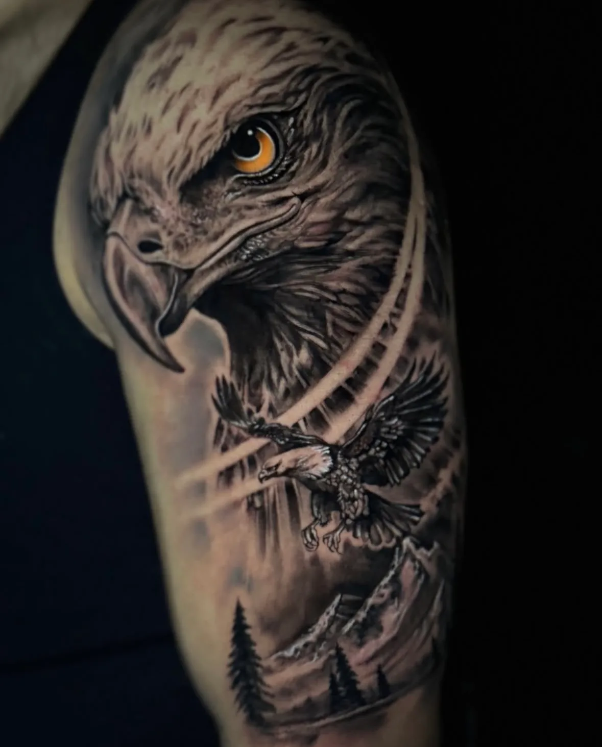 Realistisches Adler-Tattoo am Oberarm: Adlerkopf mit Bernsteinauge, fliegender Adler, Berge und Tannen, Tattoo Wien
