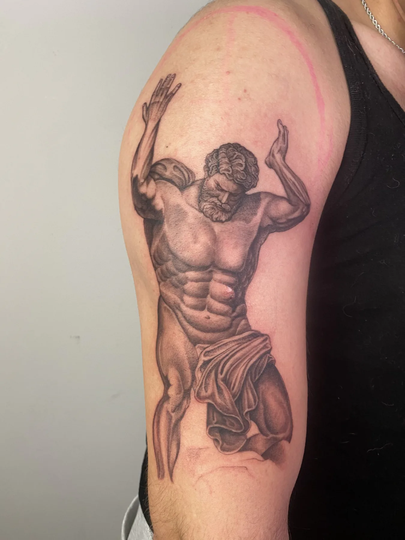 Realistisches Black & Grey Tattoo einer antiken Atlas-Statue mit erhobenen Armen und Tuch am Oberarm, Tattoo Wien