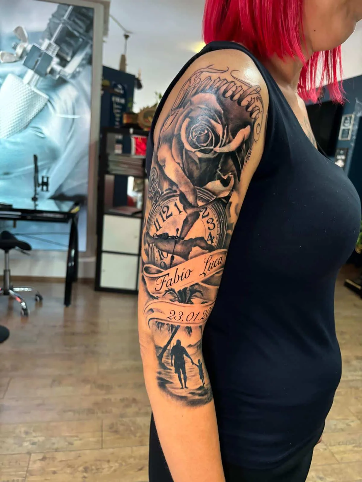 Black & Grey Half Sleeve am Oberarm mit Rose, Uhr, Namensbanner und Silhouette von Vater und Kind am Strand, Tattoo Wien