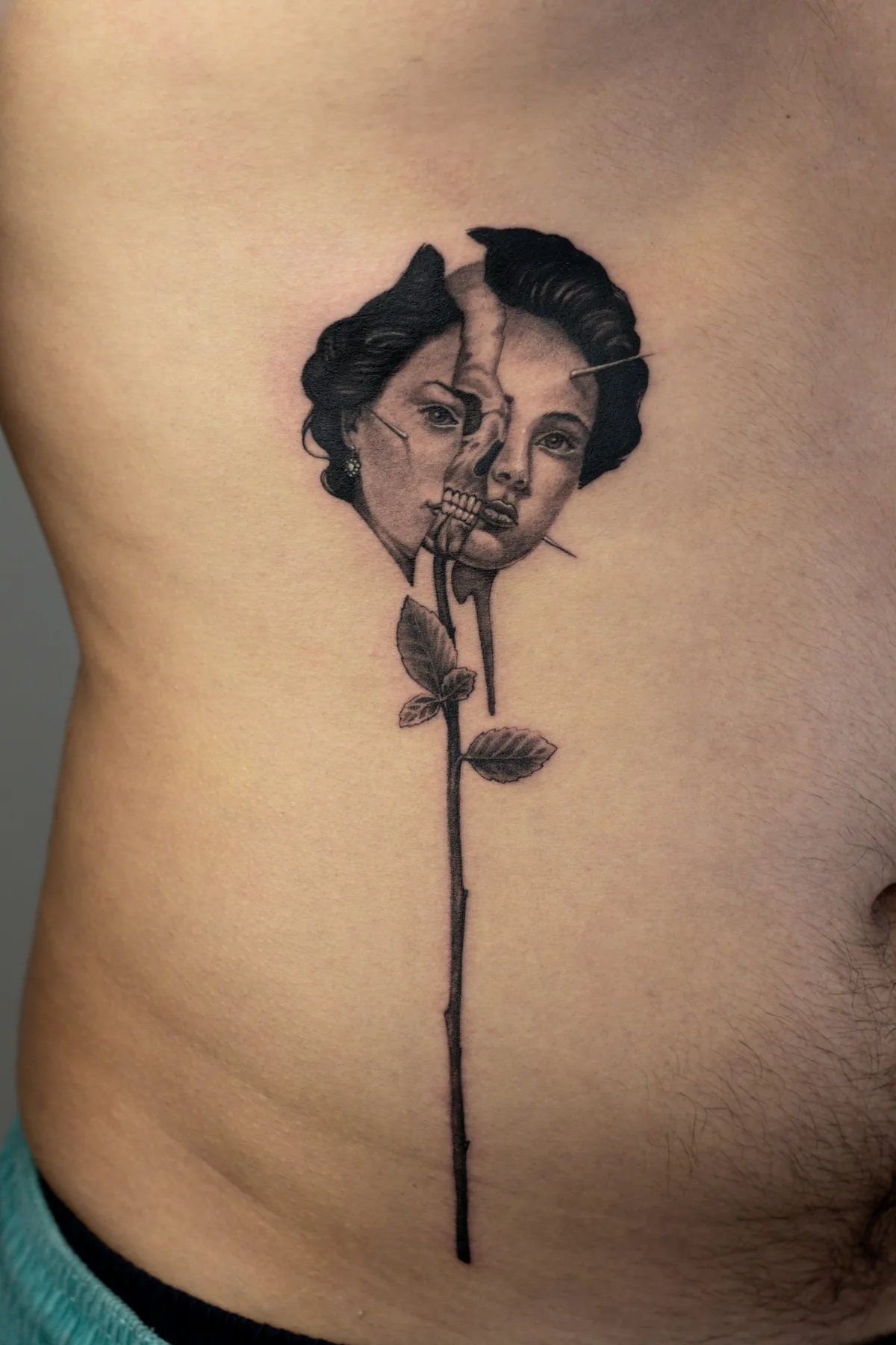 Feines Black & Grey Tattoo am Rippenbogen: gespaltenes Frauengesicht mit Totenkopf darin, auf dem langen Stiel einer Rose