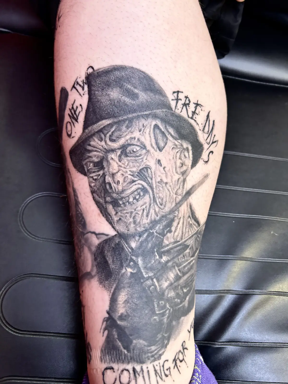 Realistisches Black & Grey Porträt-Tattoo von Freddy Krueger mit Hut und Klingenhand an der Wade, mit Schriftzug, Wien