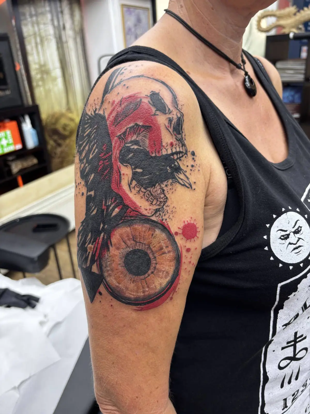 Trash Polka Tattoo an der Schulter: Rabe und Totenkopf in Schwarz mit roten Spritzern und realistischem Auge, Wien